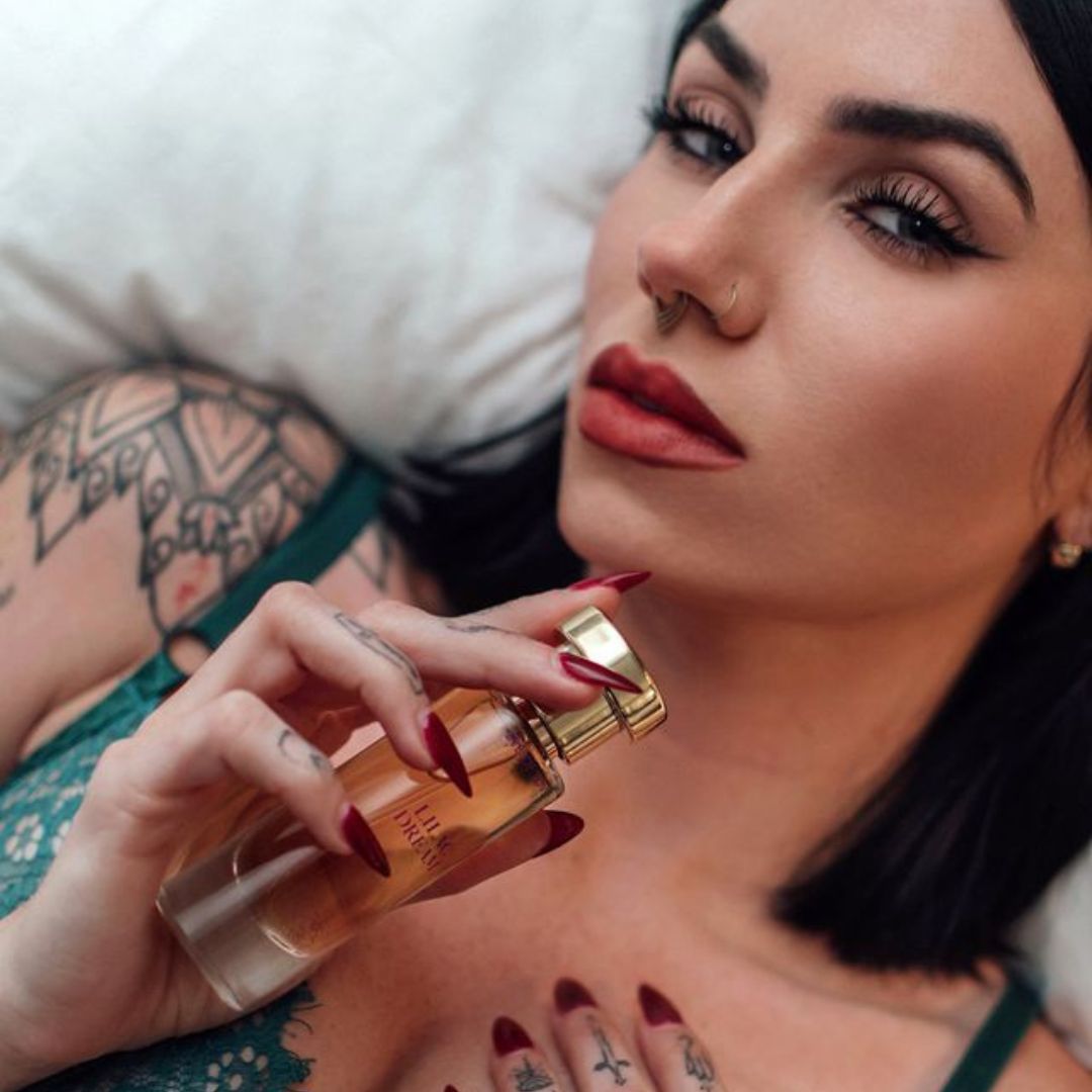 A tattooed woman with red-painted nails holding a bottle of Lilac Dreams pheromone perfume from Eye of Love.