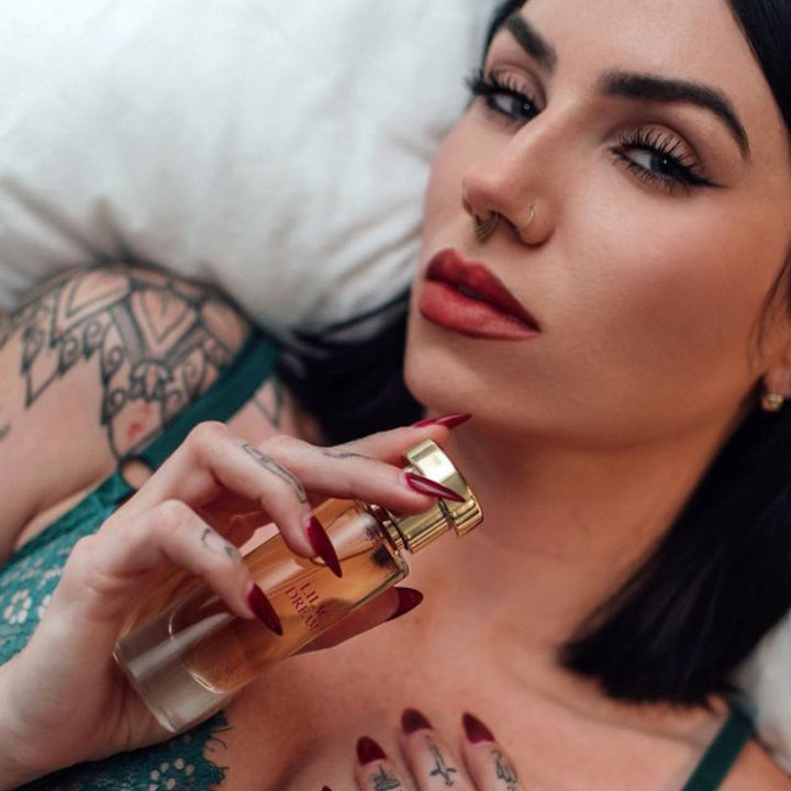 A tattooed woman with red-painted nails holding a bottle of Lilac Dreams pheromone perfume from Eye of Love.