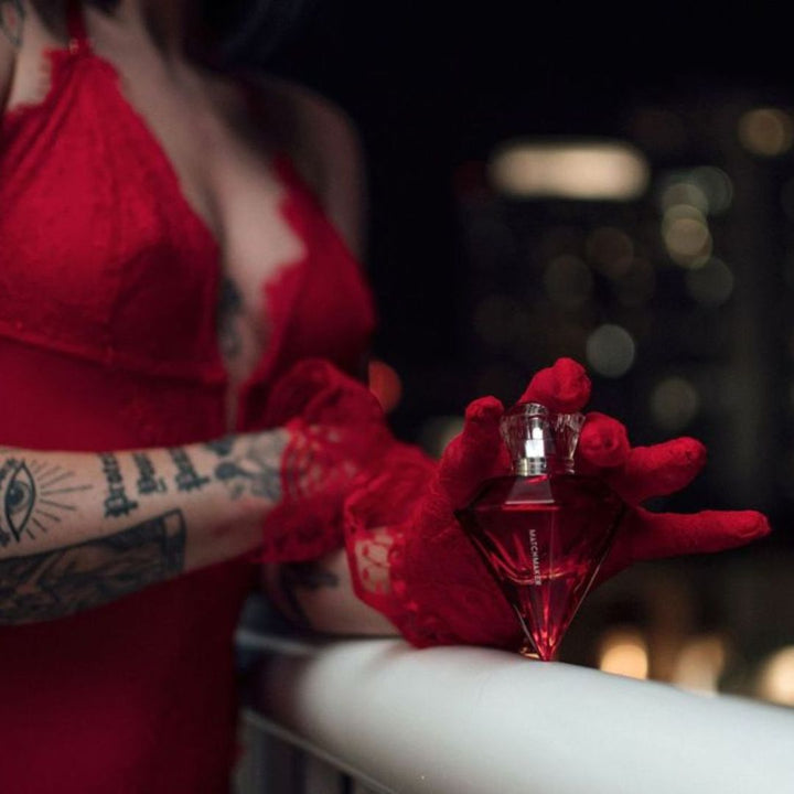 A sultry woman in a red bodysuit and lace gloves, adorned with tattoos, standing beside a white balcony rail, holding a bottle of Matchmaker pheromone perfume