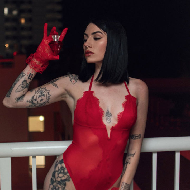 A sultry woman in a red bodysuit and lace gloves, adorned with tattoos, standing beside a white balcony rail, holding a bottle of Matchmaker pheromone perfume at eye level.