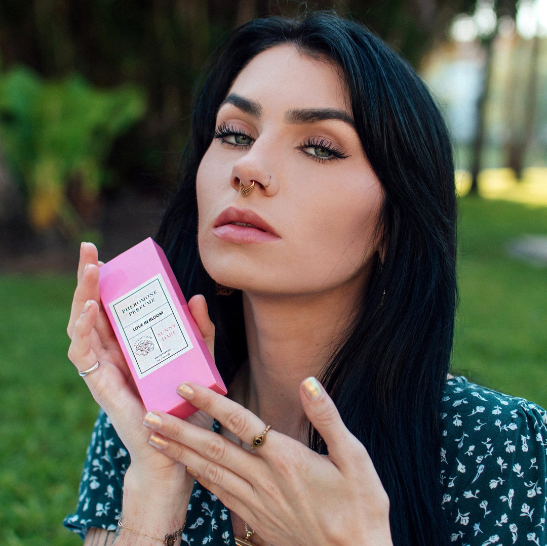 A sultry woman staring at the camera, holding the Sunny Daze pheromone perfume from Eye of Love in its box to her face.